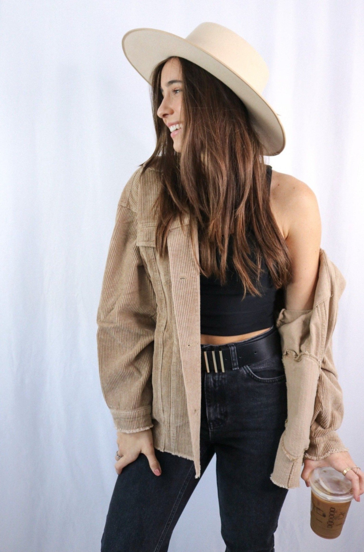 Brunette girl models buttoned corduroy jacket in the color tan for Scarlette The Label, an online fashion boutique for women. The corduroy jacket is an oversized street style jacket. Paired with a black crop top, dark denim jeans, and a wide-brimmed rancher hat.