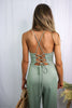 Load image into Gallery viewer, Blonde girl models crossback cami jumpsuit in color sage for Scarlette The Label, an online fashion boutique for women. Paired with a large straw bag and nude heels. 