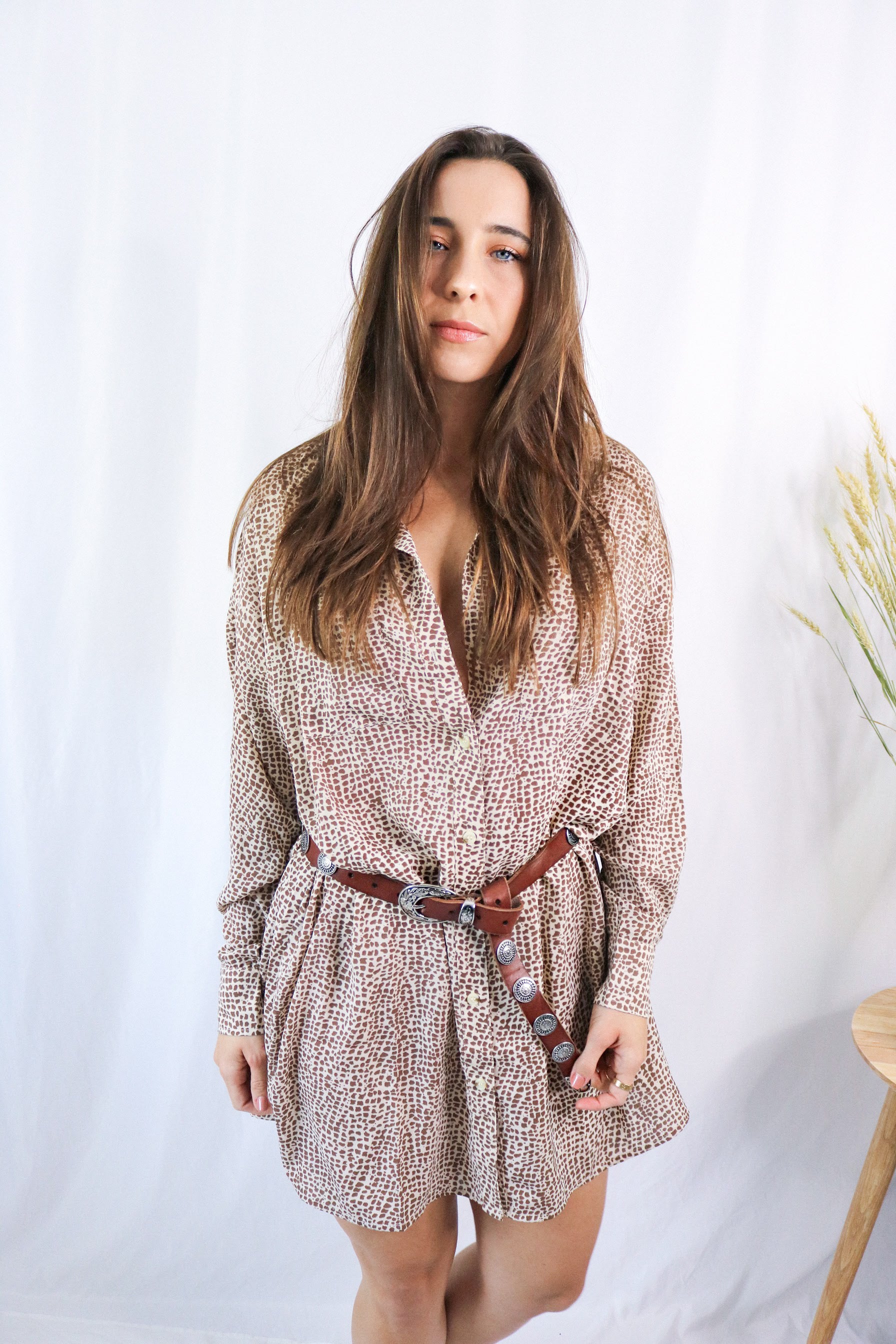 Brunette girl models a collared T-Shirt Dress in Animal Print (cheetah / leopard print). The t-shirt dress is collared, brown, and mid-length. It is a long sleeve t-shirt dress with a loose and comfortable fit and paired with a brown belt.