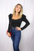 Blonde girl models a long sleeve bodysuit in black for Scarlette The Label, an online fashion boutique for women. The black bodysuit has puff sleeves. Paired with dark denim jeans and a red and gold purse clutch.