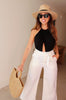 Black halter keyhole openback bodysuit from Scarlette The Label, an online fashion boutique for women. Paired with white pants, a large straw handbag and a wide brimmed hat.