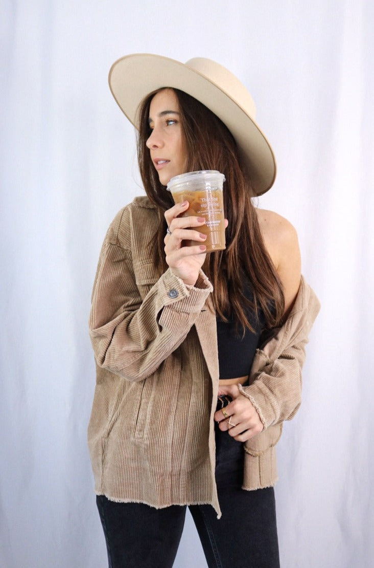 Brunette girl models buttoned corduroy jacket in the color tan for Scarlette The Label, an online fashion boutique for women. The corduroy jacket is an oversized street style jacket. Paired with a black crop top, dark denim jeans, and a wide-brimmed rancher hat.