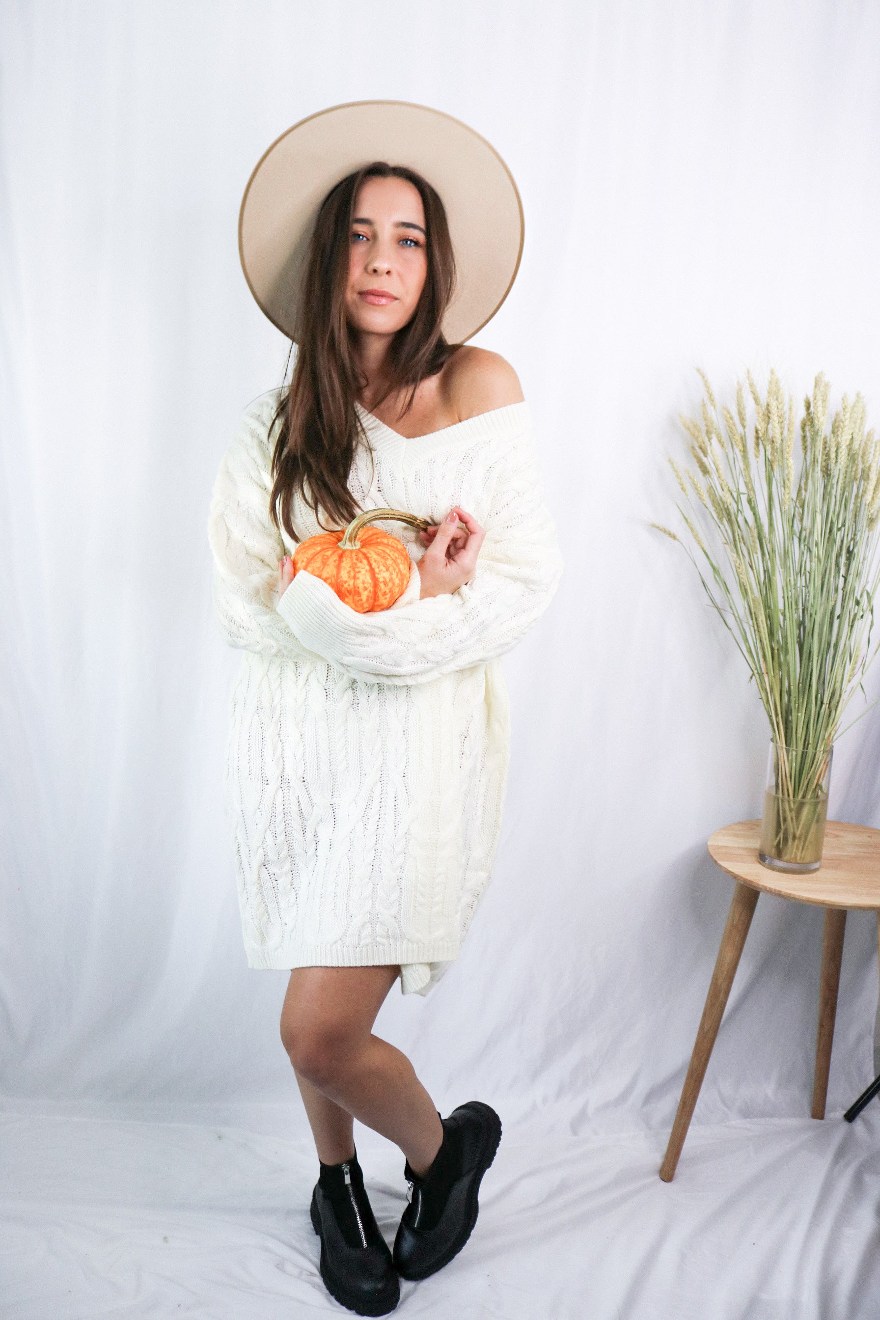 Brunette girl models a mid-length cable knit sweater dress in the color ivory for Scarlette The Label, an online fashion boutique for women. Paired with a beige wide-brimmed rancher hat. and black boots
