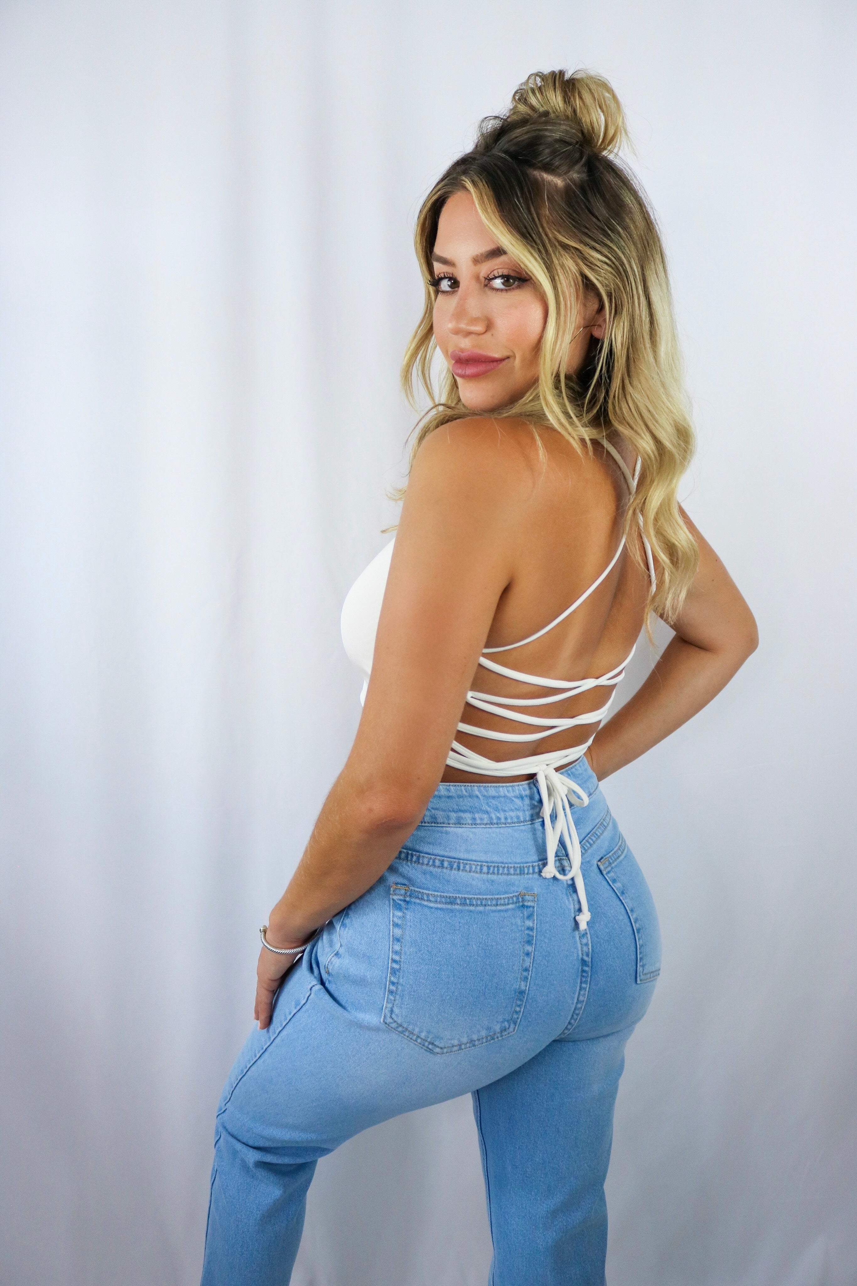 Blonde girl models a white crop top for Scarlette The Label, an online fashion boutique for women. The white crop has criss cross (crossback) detail in the back and has adjustable strings to tighten and loosen the fit. Paired with light wash denim boyfriend jeans. 
