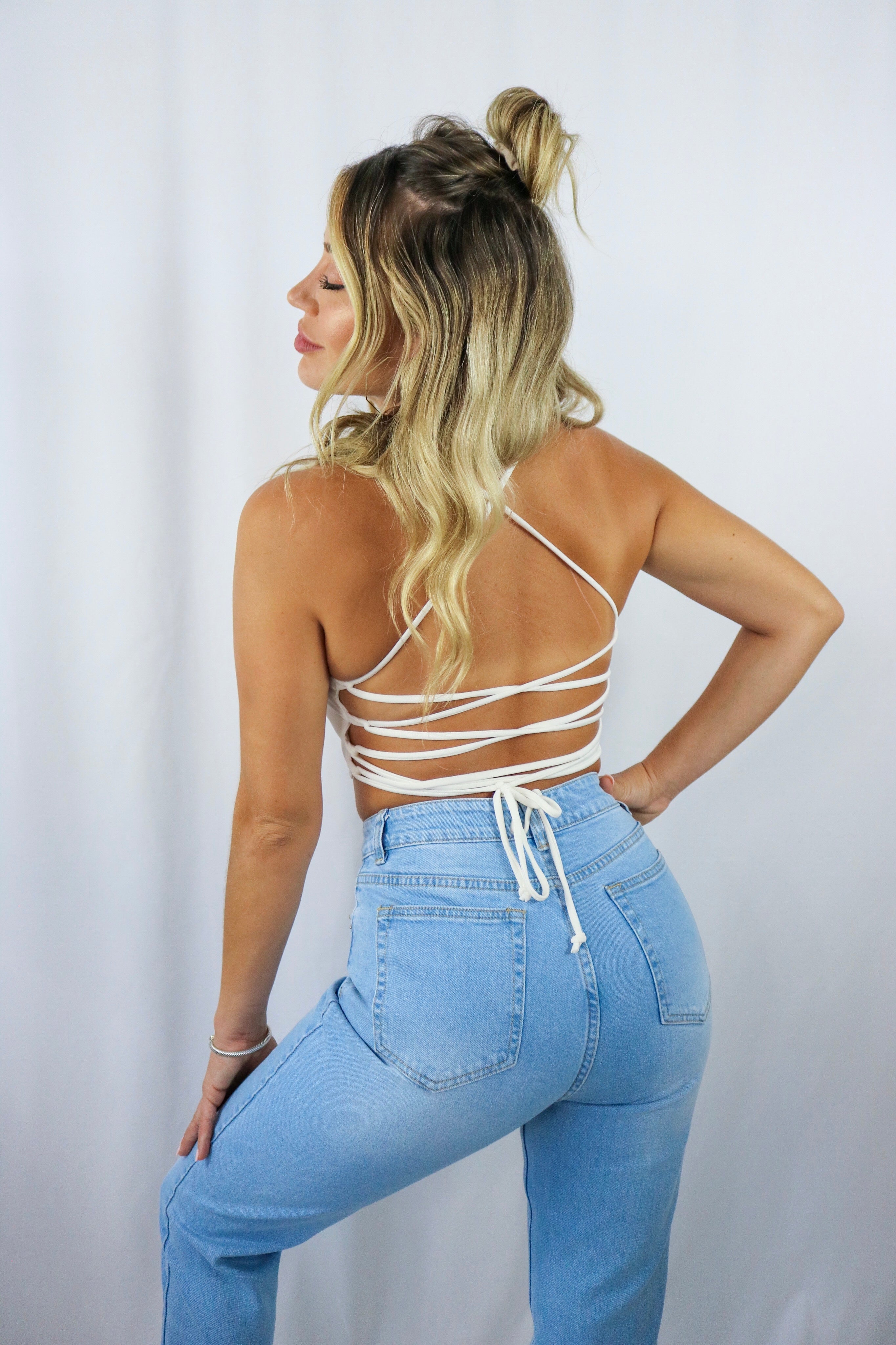Blonde girl models a white crop top for Scarlette The Label, an online fashion boutique for women. The white crop has criss cross (crossback) detail in the back and has adjustable strings to tighten and loosen the fit. Paired with light wash denim boyfriend jeans. 