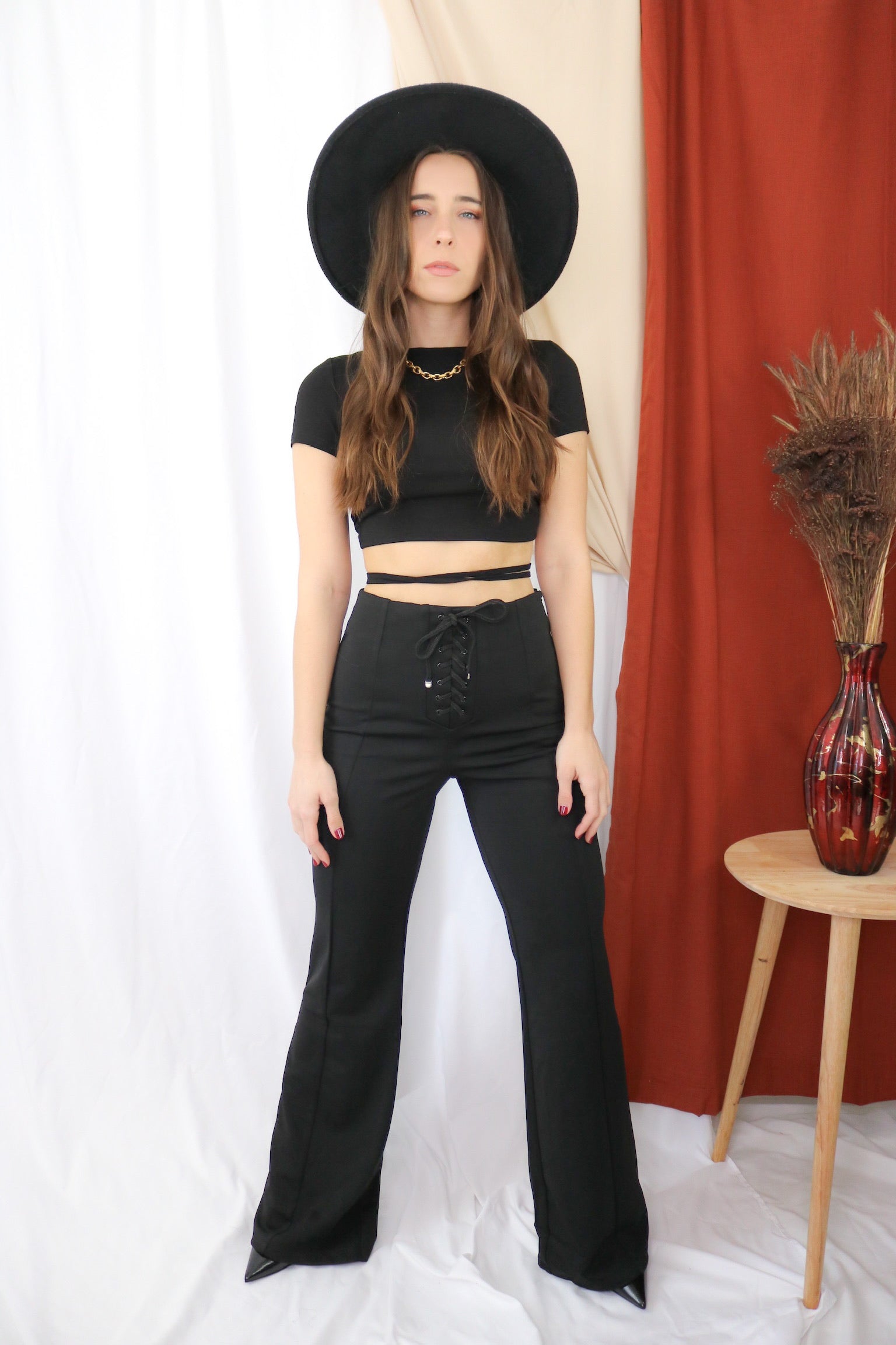 Brunette models outfit for Scarlette the Label, an online fashion boutique for women. Wears an open back black crop top and flare wide leg tie pants in black. Paired with wide brim rancher hat.