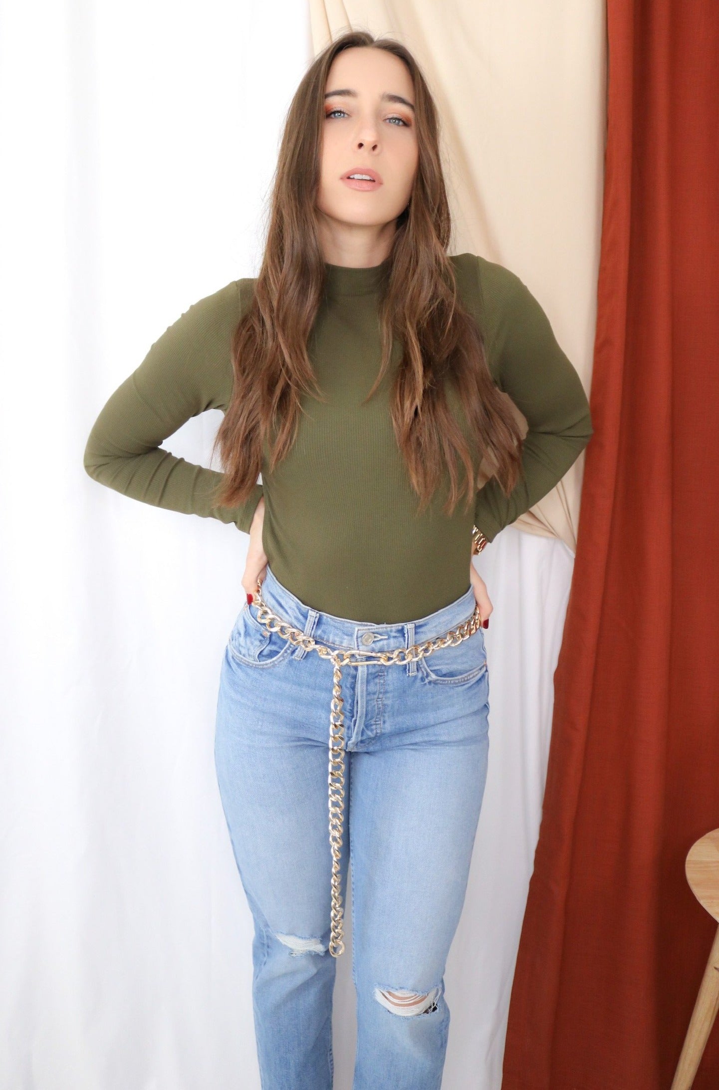 Brunette models long sleeve openback tie bodysuit in color olive for Scarlette The Label, an online fashion boutique for women. Paired with lightwash denim jeans and a gold chain belt. 