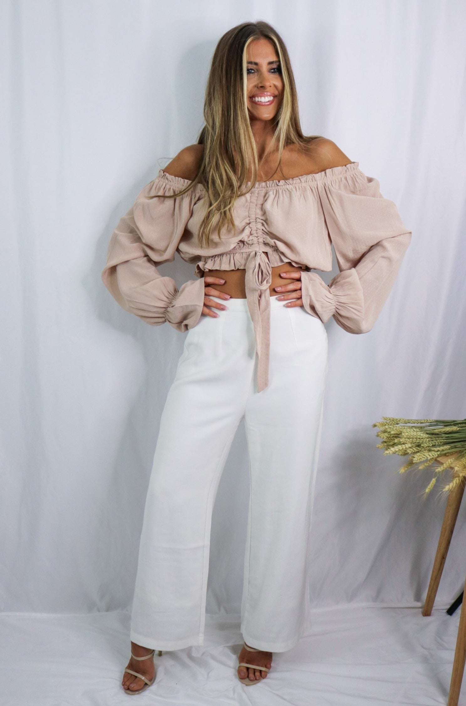 Blonde girl models an off the shoulder frilled blouse in color blush for Scarlette The Label, an online fashion boutique for women. Paired with white, wide-leg pants.