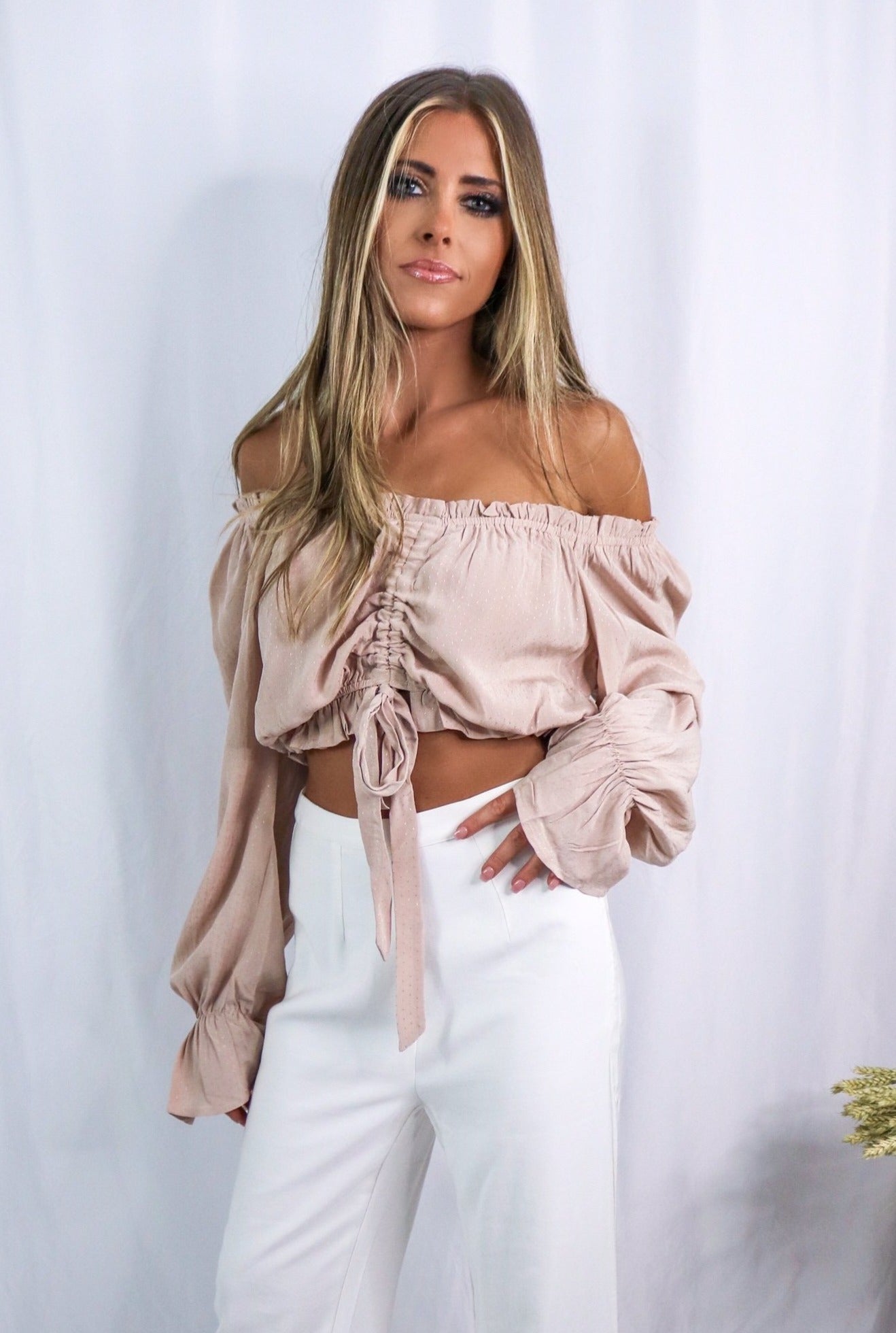 Blonde girl models an off the shoulder frilled blouse in color blush for Scarlette The Label, an online fashion boutique for women. Paired with white, wide-leg pants.