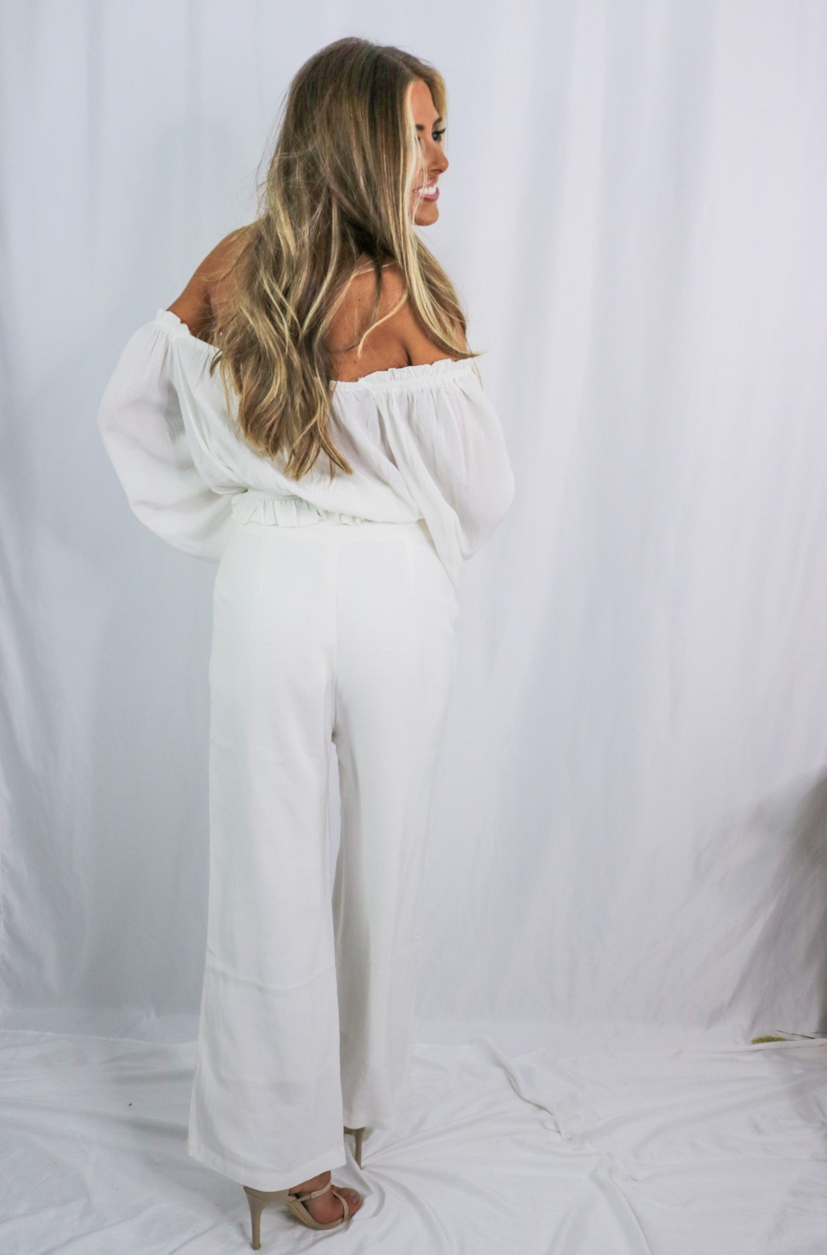 Blonde girl models an off the shoulder frilled blouse in white for Scarlette The Label, an online fashion boutique for women. Paired with white, wide-leg pants.