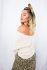 Blonde girl modeling an off the shoulder , twist front sweater in the color cream for Scarlette The Label, an online fashion boutique for women. Paired with a midi leopard maxi skirt.