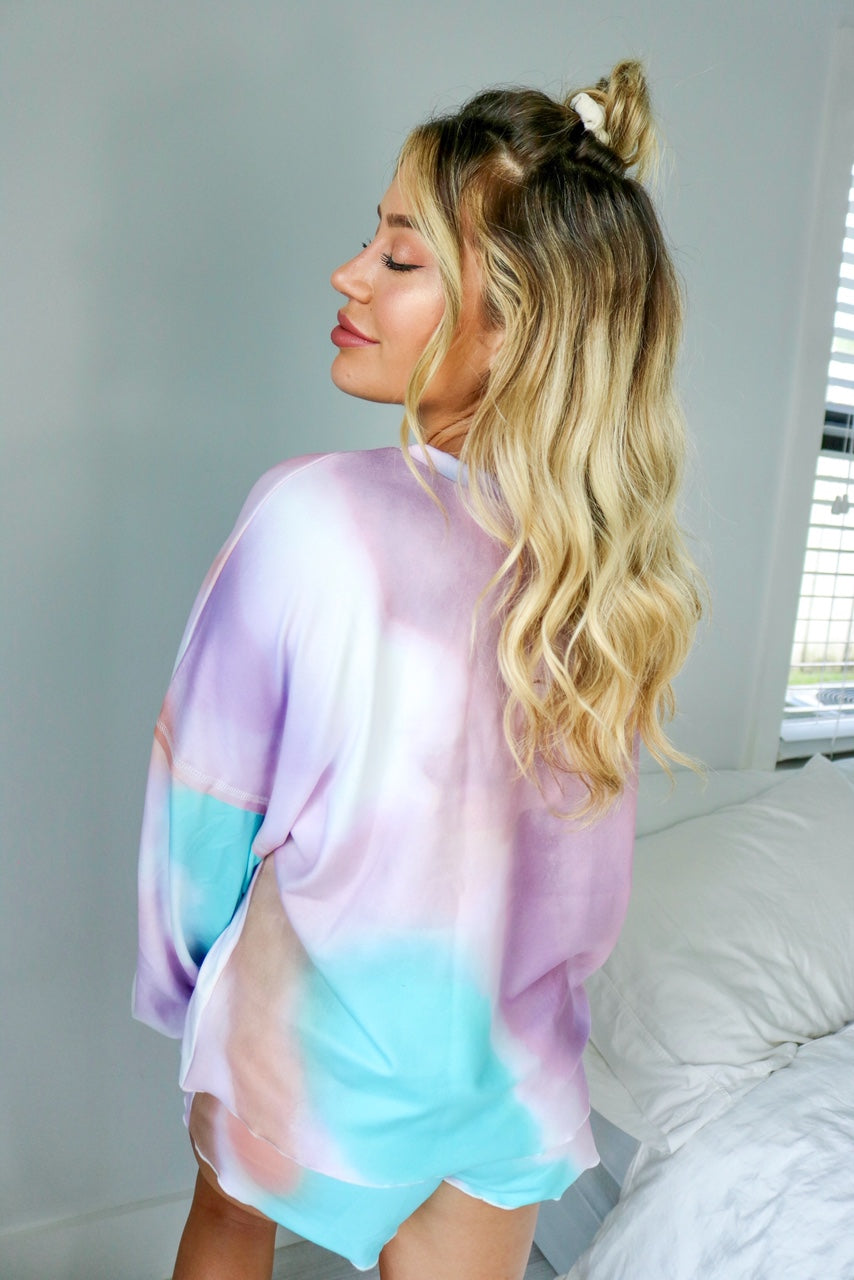 Blonde girl models a tie dye loungewear short set for Scarlette The Label, an online fashion boutique for women. The loungewear set is light blue, light purple and light pink and is a tie dye pattern. The matching tie-dye loungewear set includes a long sleeve shirt and shorts.