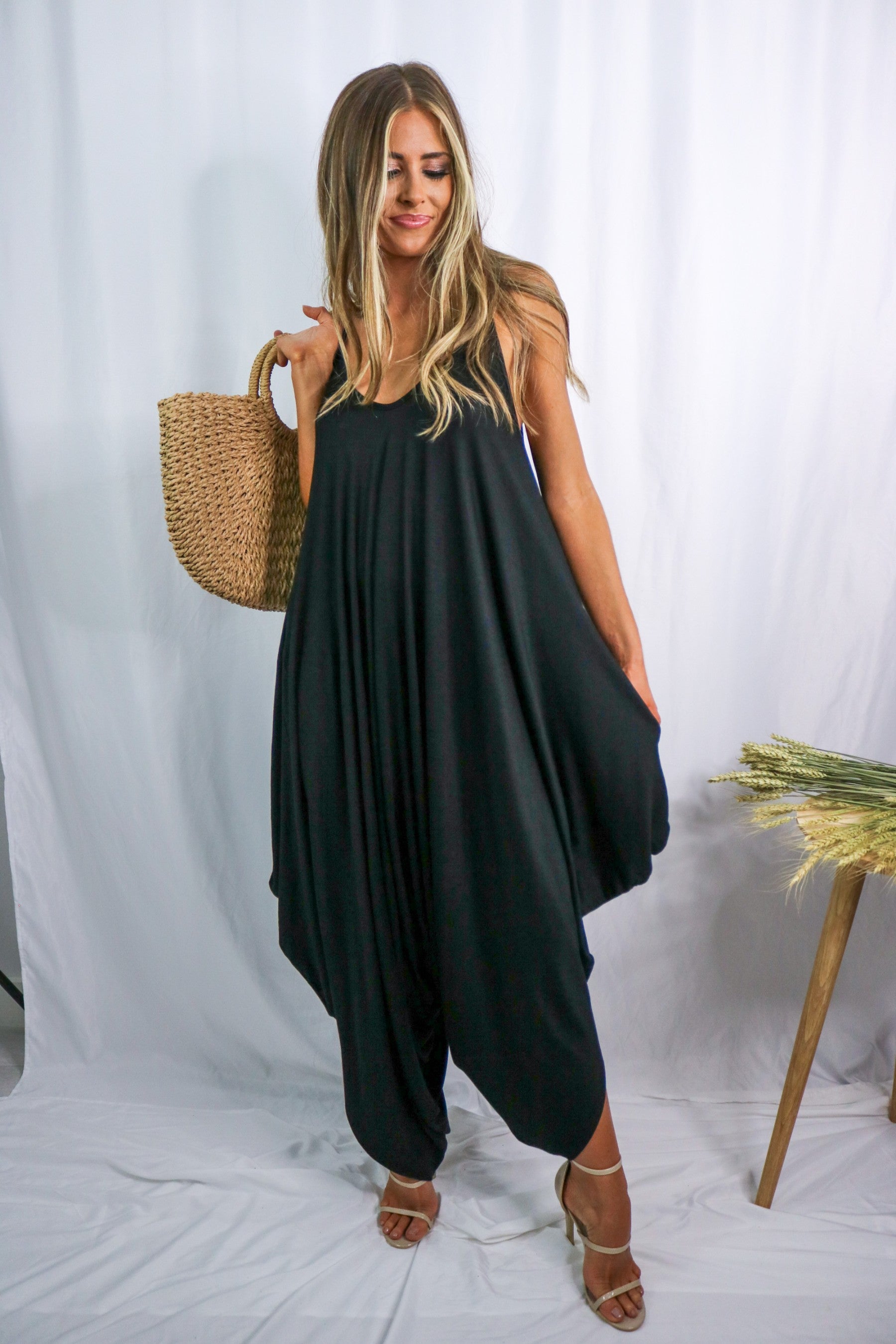 Blonde girl modeling a black boxy hi lo jumpsuit for Scarlette The Label, an online fashion boutique for women. Black jumpsuit has pockets and is lightweight and flowy. Paired with large straw bag.
