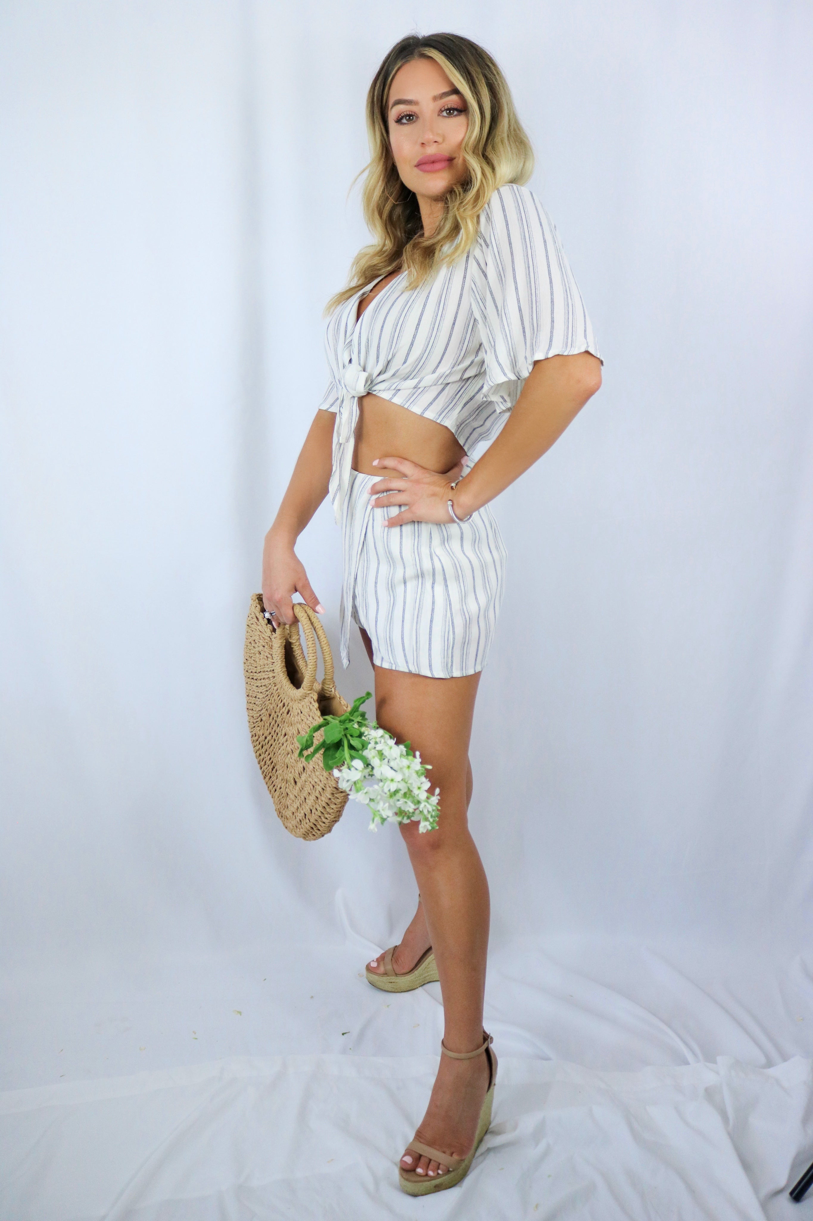 Blonde girl models a matching set for Scarlette The Label, an online fashion boutique for women. The set is a matching short set in white and charcoal stripes. Includes a short sleeve tie top and a skort. Paired with large straw bag.