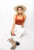 Blonde girl models a plain ponte bodysuit in the color rust for Scarlette The Label, an online fashion boutique for women. Paired with white wide-leg pants and a beige wide-brimmed rancher hat.