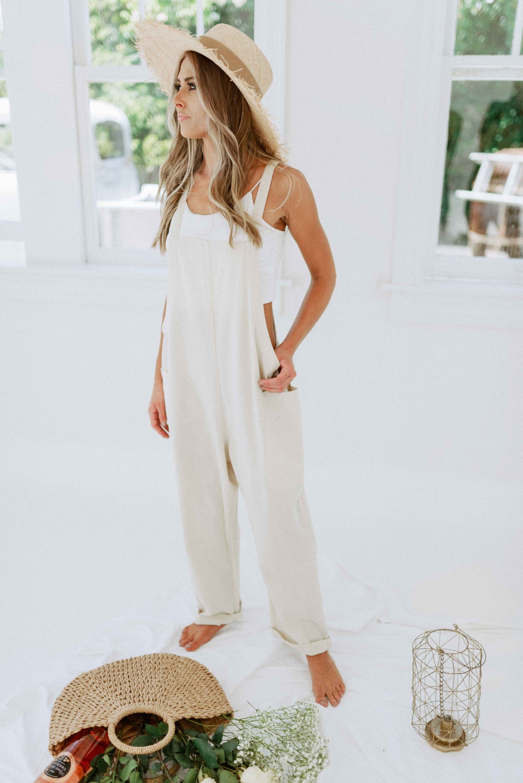 Blonde girl modeling pocketed overalls in color oatmeal for Scarlette The Label, an online fashion boutique for women. The overalls are light beige and have open sides. Paired with a wide-brimmed straw hat.