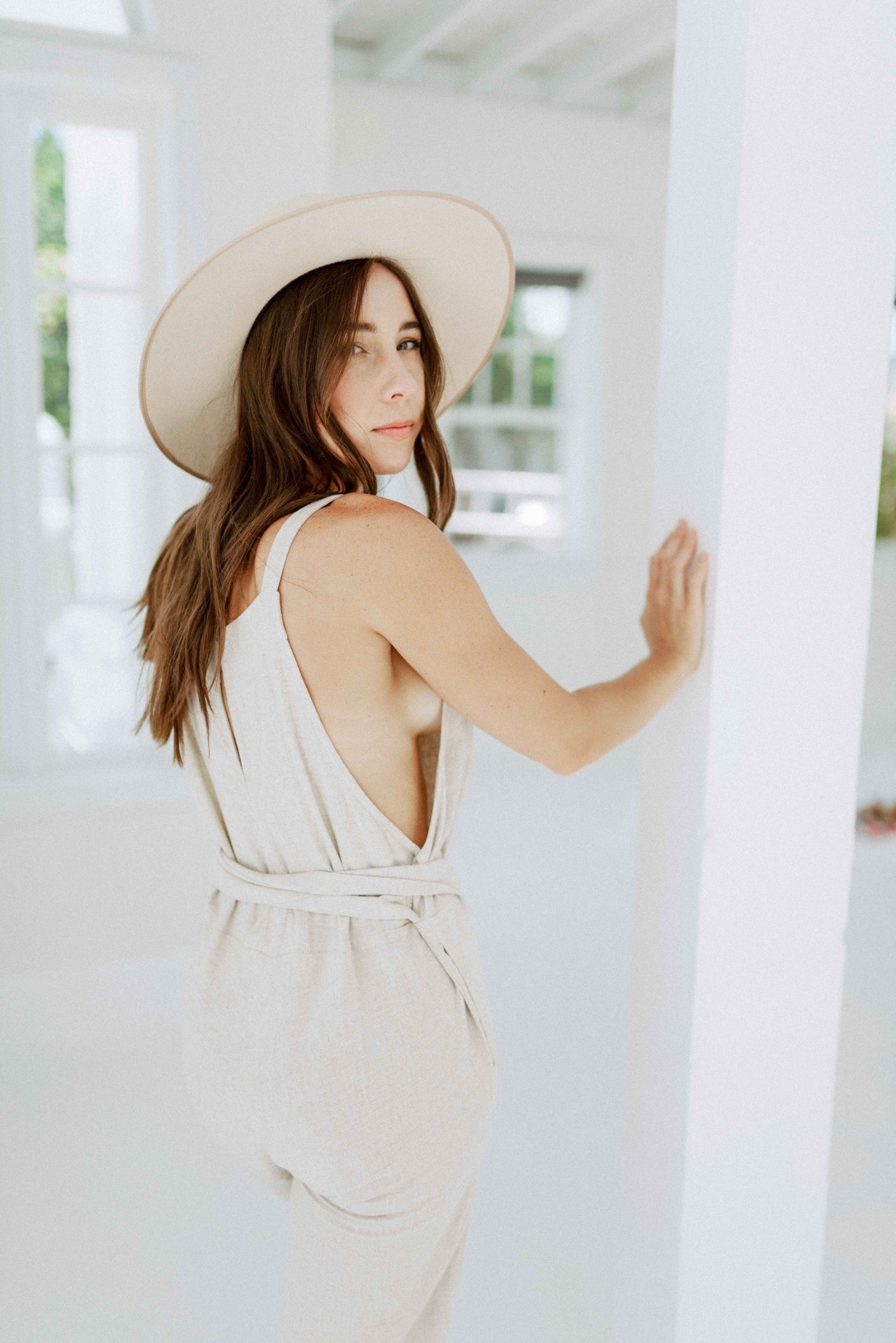 Brunette girl models beige tie jumpsuit for Scarlette The Label, an online fashion boutique for women. Paired with a beige wide-brimmed rancher hat.