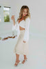 Blonde girl models ivory satin skirt and white flared sleeve tie top for Scarlette The Label, an online fashion boutique for women. The top is a white flared sleeve tie top and the skirt is a mid-length ivory satin skirt with a stretchy waist band. Paired with white heels.