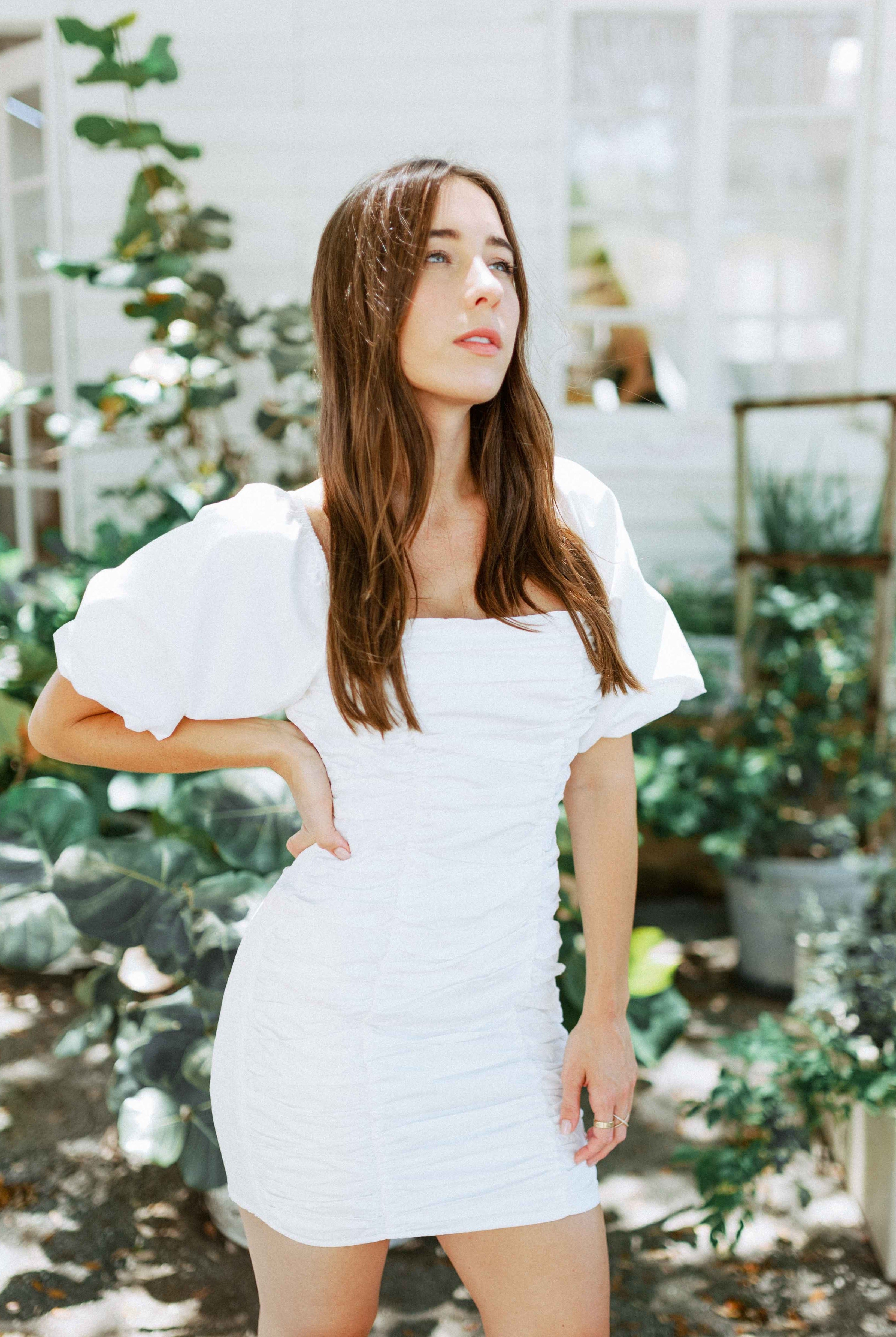 Brunette girl models white ruched mini dress with puff sleeves for Scarlette The Label, an online fashion boutique for women. White mini dress can be worn off the shoulders or regular.