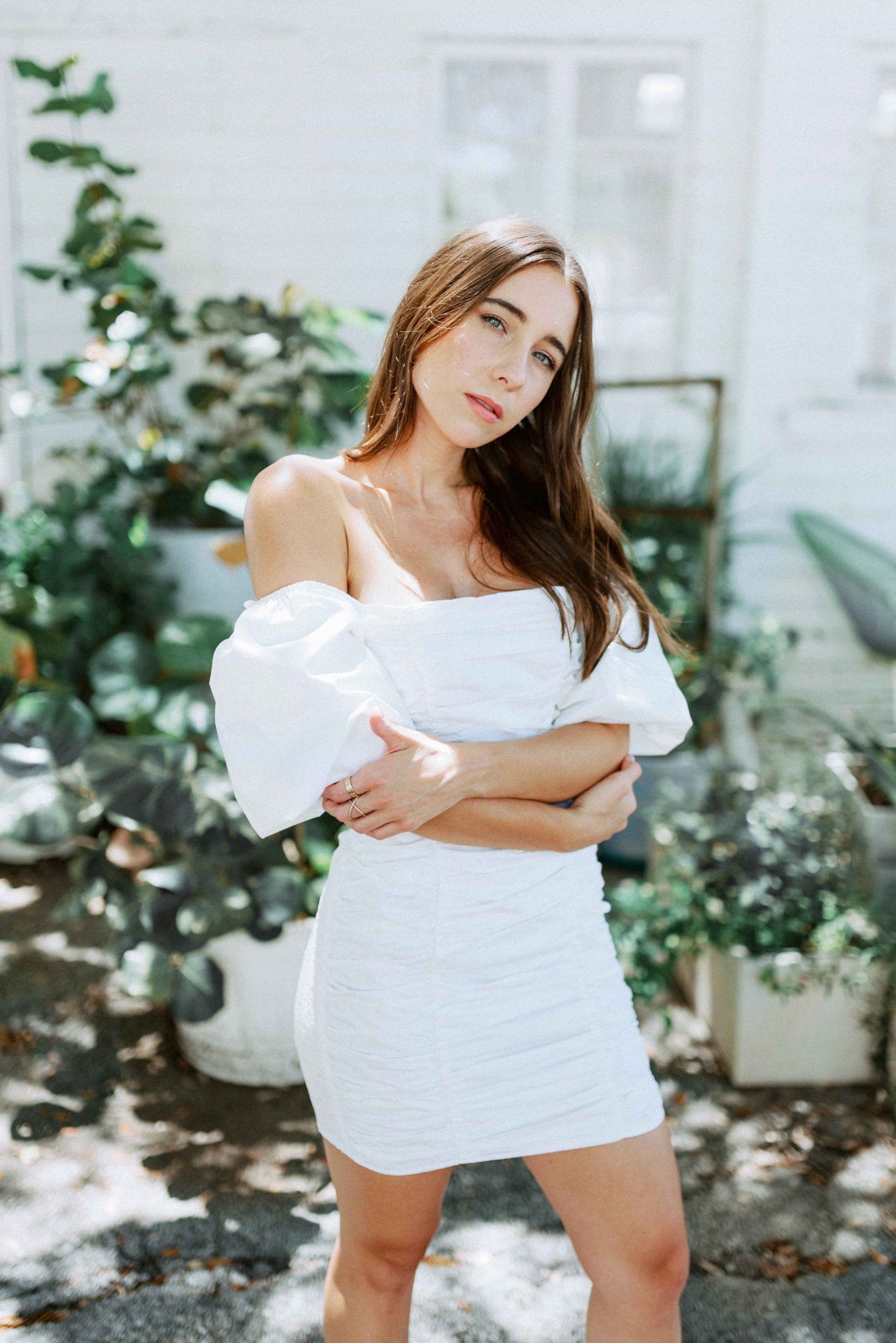 Brunette girl models white ruched mini dress with puff sleeves for Scarlette The Label, an online fashion boutique for women. White mini dress can be worn off the shoulders or regular.
