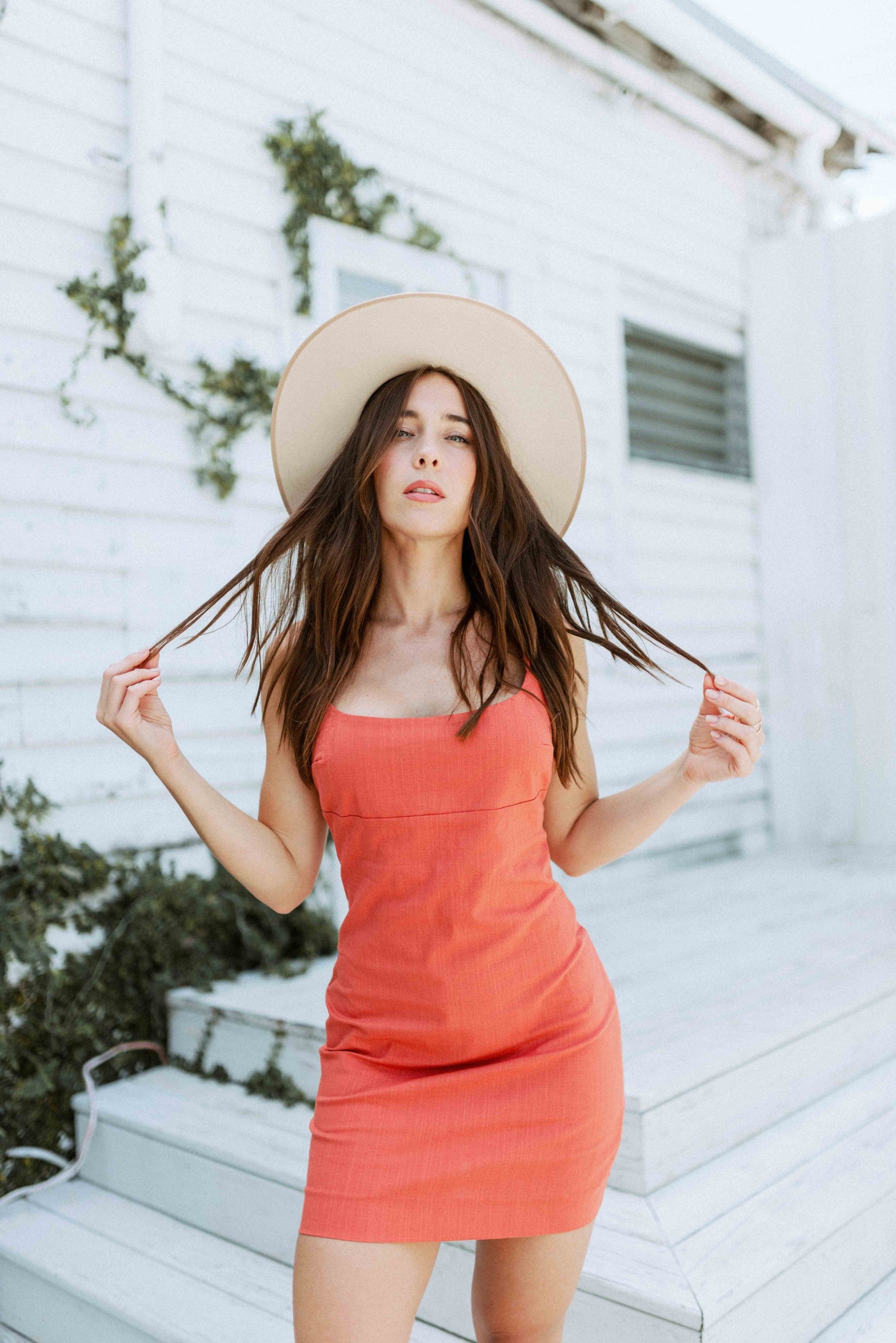 Brunette girl models a solid orange spaghetti strap mini dress for Scarlette The Label, an online fashion boutique for women. The orange mini dress is paired with a wide-brimmed rancher hat.
