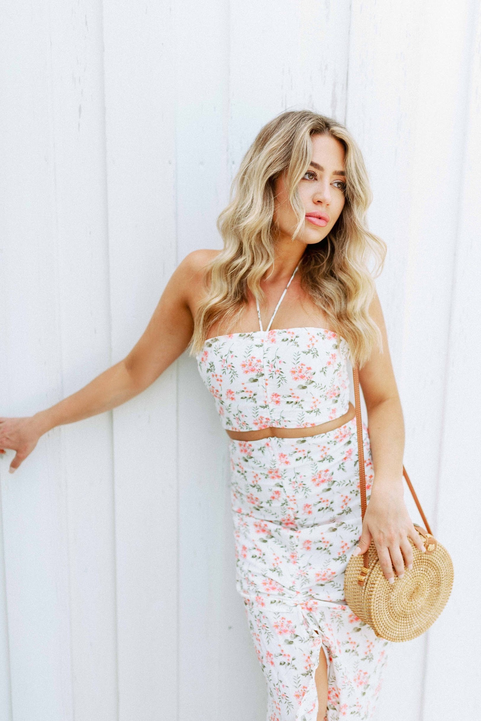 Blonde girl models a floral maxi skirt matching set for Scarlette The Label, an online fashion boutique for women.  The matching skirt set has a halter crop top and a long slitted maxi skirt. Sold separately. Paired with straw purse.